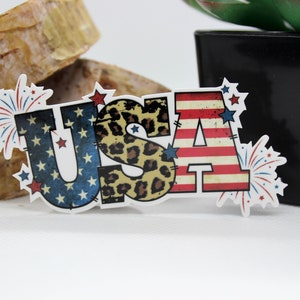 May include: A white sticker with the word "USA" in red, white, and blue letters. The letters are decorated with stars and stripes, leopard print, and fireworks.