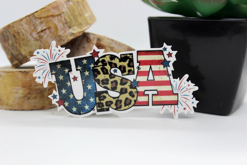 May include: A white sticker with the word "USA" in red, white, and blue letters. The letters are decorated with stars and stripes, leopard print, and fireworks.