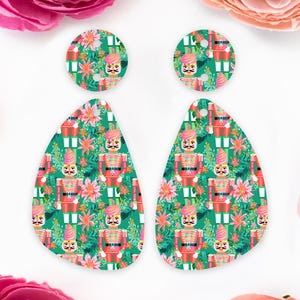 May include: Earrings with a repeating pattern of toy soldiers and floral elements on a teal background. The earrings have a teardrop shape and a circular top, suitable for festive occasions. The design is vibrant and eye-catching.