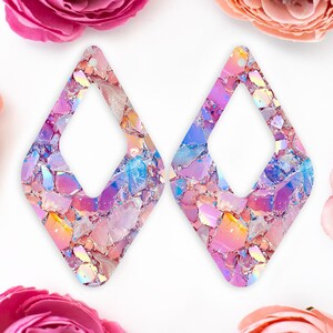 May include: A pair of iridescent pink and blue diamond-shaped earrings with a glittery finish. The earrings have a large open space in the centre.