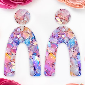 May include: A pair of iridescent, arch-shaped earrings with a circular top. The earrings are made of a mosaic of colourful, reflective pieces in shades of pink, purple, blue, and yellow. The earrings are set against a white background with pink flowers.