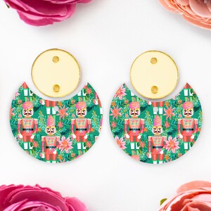 May include: A pair of festive earrings. Each earring features a gold-coloured top with two holes and a half-moon shaped bottom. The bottom has a green background with a repeating pattern of toy soldiers and floral accents.