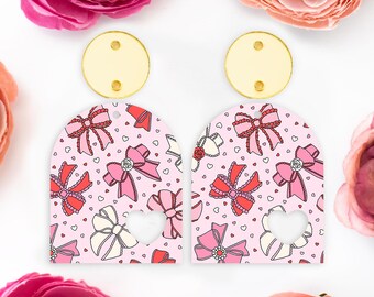 May include: A pair of earrings with a pink bow pattern and a heart cutout. The earrings have a gold-colored top piece. The design includes bows, hearts, and roses on a pink background.