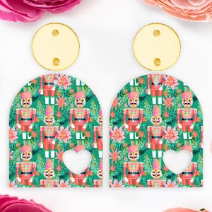 May include: A pair of festive earrings with a green background patterned with red and gold nutcrackers and pink flowers. Each earring has a gold circle top and a heart-shaped cutout.