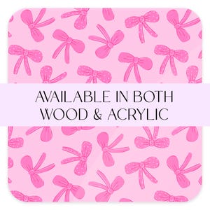 May include: A pink background with a pattern of pink bows, some with white polka dots. The text "AVAILABLE IN BOTH WOOD & ACRYLIC" is displayed in a white banner across the image.