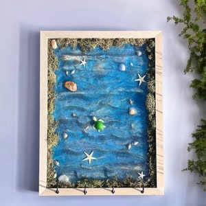 May include: A framed artwork depicting a blue ocean scene with a small green turtle, seashells, and starfish. The frame is made of wood and has a light brown finish. There are three hooks on the bottom of the frame.