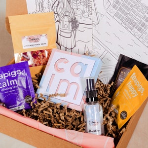 May include: A cardboard box filled with a self-care gift set. The box contains tea bags, bath salts, a small bottle of essential oil, and a card that says "calm".