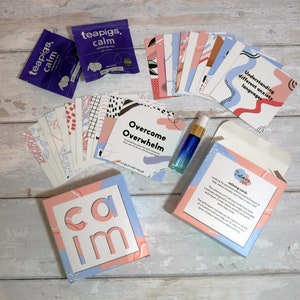May include: A set of calming cards in a pink and blue box with the word "calm" on the front. The box is surrounded by a variety of items, including tea bags, a small bottle of essential oil, and a set of cards with different designs. The cards are designed to help reduce anxiety and stress.