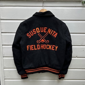 Peut inclure: Veste de baseball noire avec des poignets et une ceinture rayés orange et noirs. Le dos de la veste présente un écusson brodé avec le texte "Susquenita Field Hockey" et deux crosses de hockey sur gazon croisées.