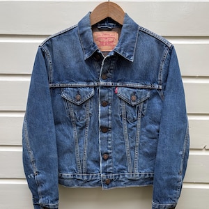 Op de afbeelding: Een blauwe spijkerjas met knoopsluiting, twee borstzakken en een rood label. De jas hangt aan een houten hanger. De jas heeft een vintage uitstraling. De achtergrond is wit.