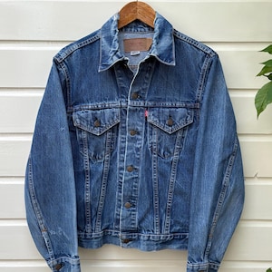 Op de afbeelding: Een medium-wash blauwe spijkerjas met een puntige kraag, lange mouwen met knoopmanchetten en een knoopsluiting aan de voorzijde. De jas heeft twee borstzakken met knoopkleppen, een klein rood merklabel en een bruin leren label in de nek.