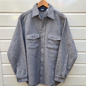 May include: A gray herringbone-patterned shirt jacket with two chest pockets and button closures. The long-sleeved shirt is hanging on a wooden hanger against a white and brown background.