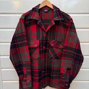 Op de afbeelding: Een rood, zwart en groen geruit flanellen overhemd met twee borstzakken en rode knopen. Het overhemd hangt aan een houten hanger tegen een witte achtergrond. Het overhemd heeft een klassieke, vintage stijl.