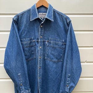 Op de afbeelding: Een blauw spijkeroverhemd met twee borstzakken en knoopsluitingen. Het shirt met lange mouwen hangt aan een houten hanger. Het shirt heeft een klassieke kraag en manchetten met knopen.