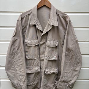 Könnte beinhalten: Eine beige Utility-Jacke mit mehreren Fronttaschen und Knopfverschlüssen. Die Jacke hängt an einem Holzbügel vor einer weiß gestrichenen Holzwand. Die Jacke hat einen Kragen und lange Ärmel.