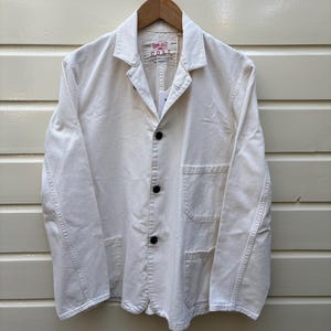 May include: Off-white work coat with three black buttons and two front pockets. The coat is hanging on a wooden hanger. A tag is attached to the coat with the text "COAT" and other text.