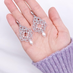 Può includere: Un paio di orecchini traforati color argento con un motivo floreale e una perla pendente. Gli orecchini sono tenuti in una mano, mostrando i loro dettagli intricati e lo stile elegante. Lo sfondo è bianco.