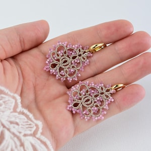 Pode incluir: Um par de brincos pendentes com um intrincado trabalho de fio dourado num padrão de renda circular, adornados com pequenas contas quadradas cor-de-rosa claro nas bordas e finalizados com fechos de alavanca em metal dourado polido.