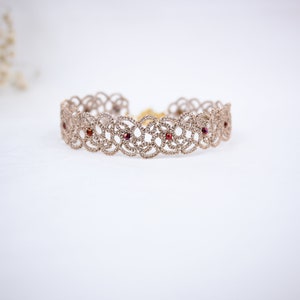 Pode incluir: Um delicado bracelete dourado com um design de renda e pequenas pedras preciosas vermelhas.