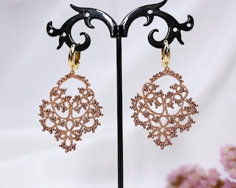 May include: A pair of rose gold hoop earrings with a delicate, intricate lace design. The earrings are hanging from a black display stand.