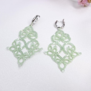 May include: A pair of delicate green dangle earrings with an intricate lace-like design. The earrings are made of small beads and have a silver hook fastening.