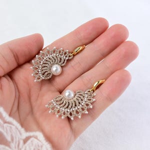 Pode incluir: Um par de brincos em forma de leque. Os brincos apresentam um design em metal dourado com detalhes intrincados, pequenas pérolas brancas e uma pérola maior no centro. São exibidos sobre um fundo branco suave.