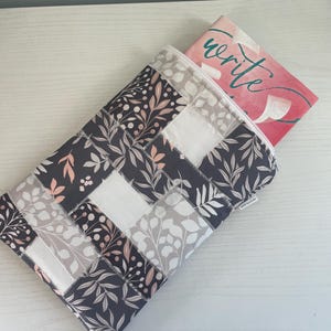 May include: A quilted book sleeve with a patchwork design. The sleeve features a pattern of gray, white, and pink floral fabric. A book with the word "write" is partially visible inside the sleeve.
