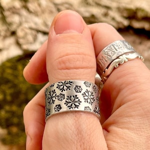 Puede incluir: Un anillo de plata con un diseño de copo de nieve. El anillo se lleva en el pulgar.