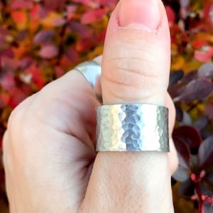 May include: A wide, hammered silver ring is worn on a finger, showcasing a textured surface. The ring's design features a series of small, irregular indentations, creating a unique, handcrafted appearance. The background is a colorful display of red and burgundy foliage.
