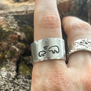 May include: A hand wearing three silver rings. The ring on the index finger features a design of two bears and a heart. The ring on the thumb has the initials "S & R" engraved on it.