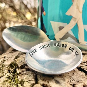 Op de afbeelding: Een zilveren lepel met de inscriptie "pour some sugar on me" op een houten oppervlak. Op de achtergrond staat een blauwe mok met een afbeelding van twee mensen die lopen.