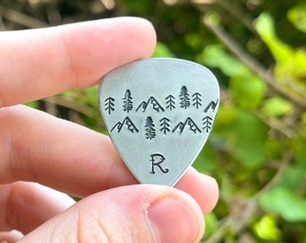 May include: A silver guitar pick with a mountain and tree design. The pick has the letter "R" stamped on it. The pick is held in front of a blurred green background.