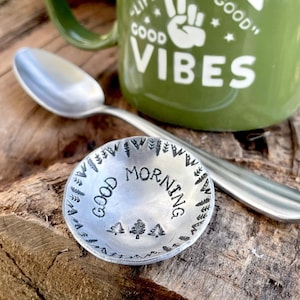 May include: A green ceramic mug with the text "Spread Good Vibes" and a peace sign. The mug is sitting on a wooden surface next to a small silver dish with the text "Good Morning" and a tree design.