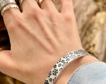 May include: A silver bracelet with a snowflake pattern. The bracelet is worn on a person's wrist.