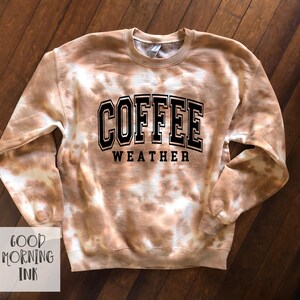 K&ouml;nnte beinhalten: Ein braun-wei&szlig;es Batik-Sweatshirt mit den Worten "COFFEE WEATHER" in schwarzen Blockbuchstaben. Das Sweatshirt hat einen Rundhalsausschnitt und lange &Auml;rmel. Die Worte "GOOD MORNING INK" stehen in einer wei&szlig;en Box unten links.