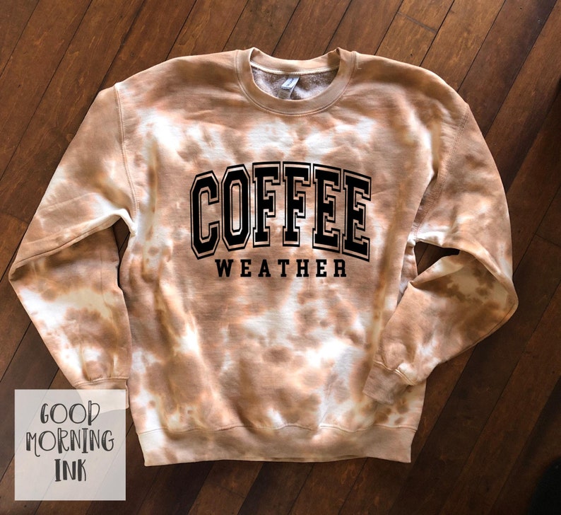 K&ouml;nnte beinhalten: Ein braun-wei&szlig;es Batik-Sweatshirt mit den Worten "COFFEE WEATHER" in schwarzen Blockbuchstaben. Das Sweatshirt hat einen Rundhalsausschnitt und lange &Auml;rmel. Die Worte "GOOD MORNING INK" stehen in einer wei&szlig;en Box unten links.