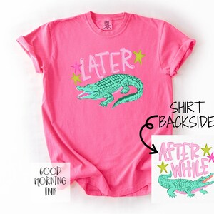 以下が含まれることがあります： 明るいピンク色のTシャツ。ライトピンクで「LATER」の文字と緑色のアリゲーターのイラストが描かれています。ピンクと黄色の2つの星も特徴です。Tシャツの裏側には「AFTER WHILE」の文字と別のアリゲーターのイラストが描かれています。