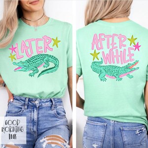以下が含まれることがあります： 漫画のアリゲーターのデザインが施された、明るい緑色のTシャツ。「LATER」の文字がピンク色で、黄色の星が描かれています。Tシャツの背面には、お揃いのアリゲーターのデザインで「AFTER WHILE」と書かれています。