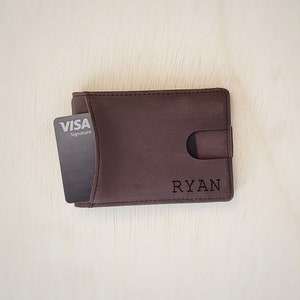 A top-down view of a slim minimalist bifold wallet laying front side up on top of a desk. The bottom front right corner shows a clean custom engraving of a name. A card is shown being slightly pushed out of the front access card slot on the front of the wallet.