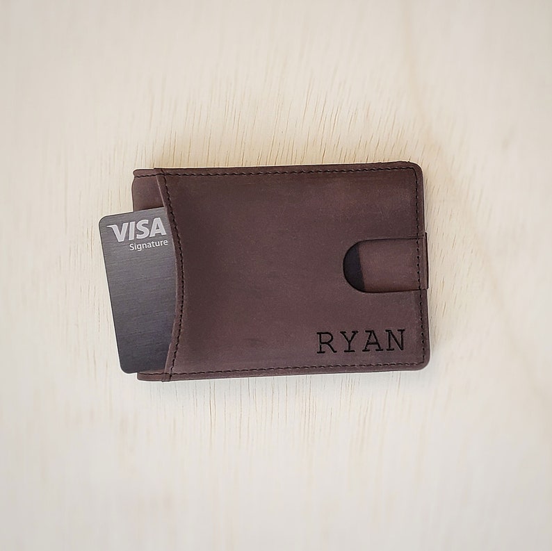 A top-down view of a slim minimalist bifold wallet laying front side up on top of a desk. The bottom front right corner shows a clean custom engraving of a name. A card is shown being slightly pushed out of the front access card slot on the front of the wallet.