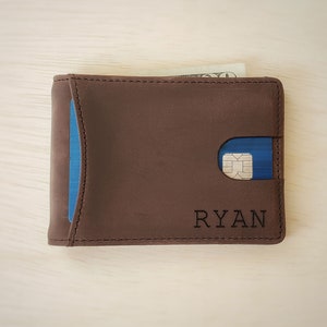 A top-down view of a slim minimalist bifold wallet laying front side up on top of a desk. The bottom front right corner shows a clean custom engraving of a name. The front of the wallet shows a card in a quick access card slot accessible by pushing the card out with the thumb.