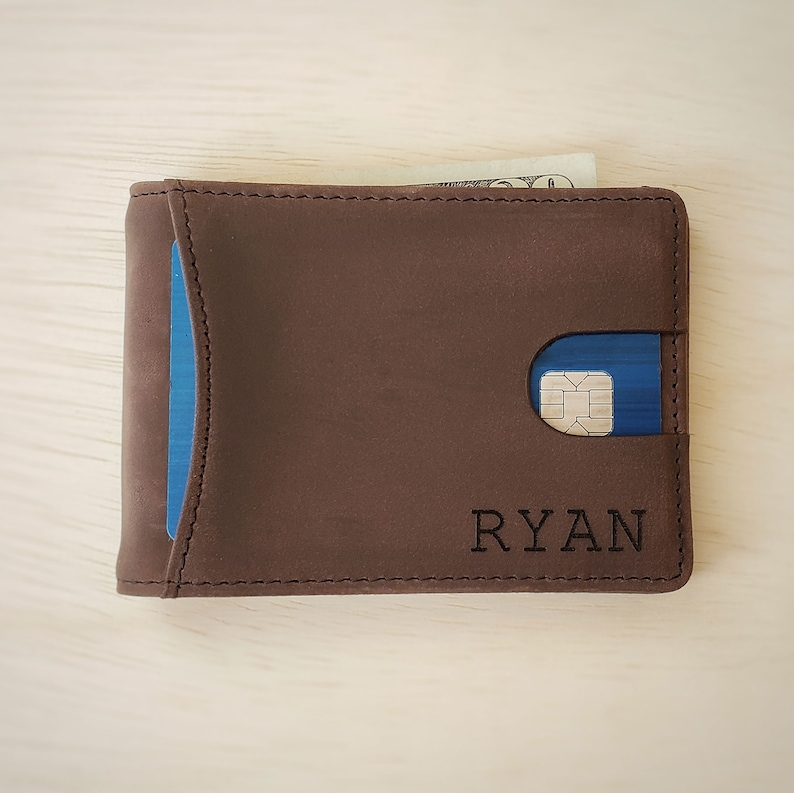 A top-down view of a slim minimalist bifold wallet laying front side up on top of a desk. The bottom front right corner shows a clean custom engraving of a name. The front of the wallet shows a card in a quick access card slot accessible by pushing the card out with the thumb.