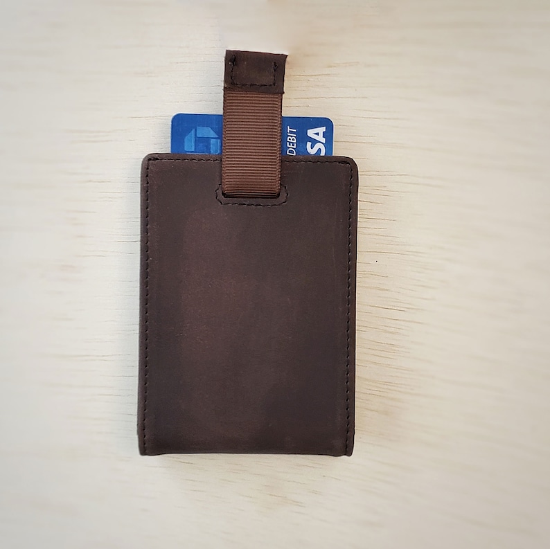 A top-down view of a slim minimalist bifold wallet laying back side down on top of a desk. The top end of the wallet shows a pull tab revealing a couple cards coming out of a card pocket.