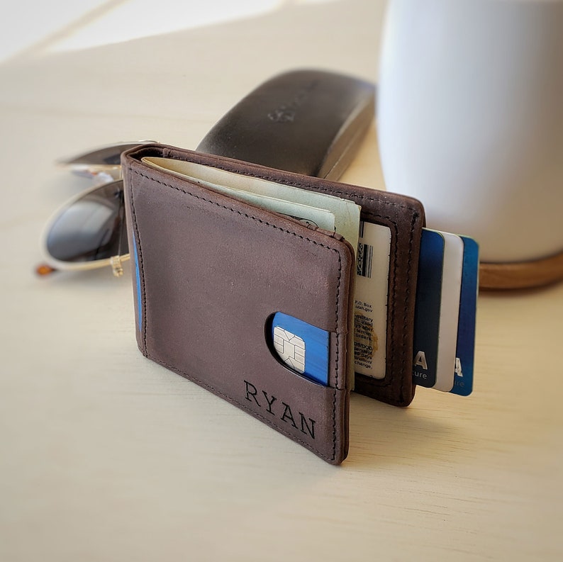 A dark brown slim leather minimalist bifold wallet stands upright on a desk, slightly open with a personalized engraving displaying a name on the front bottom right portion of the wallet. Inside the wallet some money is visible held by a money clip, and some cards hang out of the card pocket at the end of the wallet on the right side. The front side of the wallet holds a card in the quick access slot.