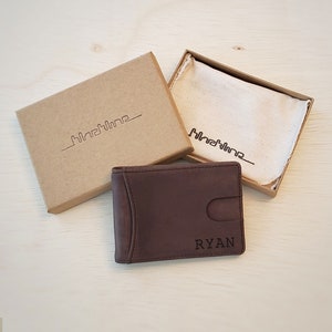A dark brown bifold genuine leather slim wallet rests on top an open wallet box for shipping. The wallet has a custom engraving on the bottom front right corner. The recycled eco-friendly box is sized to fit one slim split billfold wallet inside.