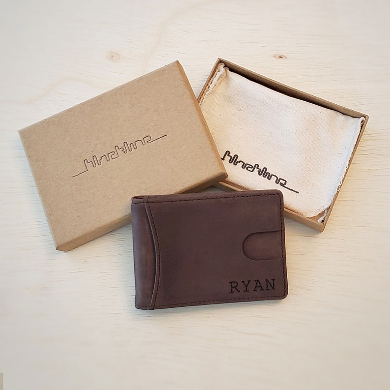 A dark brown bifold genuine leather slim wallet rests on top an open wallet box for shipping. The wallet has a custom engraving on the bottom front right corner. The recycled eco-friendly box is sized to fit one slim split billfold wallet inside.