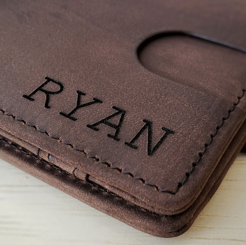 A custom name is engraved on the front right bottom area of a dark brown full grain slim leather wallet. The view is zoomed in to show the quality of the black engraving and the genuine leather. Dark brown stitching curls around the rounded corner of the wallet. A quick access card slot is visible on the front of the wallet.