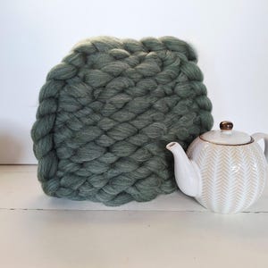 Könnte beinhalten: Ein großes, handgestricktes, salbeigrünes Kissen mit einer groben, geflochtenen Textur. Daneben steht eine weiße Keramik-Teekanne mit goldenem Deckel und Fischgrätmuster.
