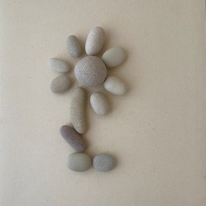 Op de afbeelding: Een bloem gemaakt van kleine, gladde, grijze stenen op een beige achtergrond. De bloem heeft een centrum gemaakt van een grotere, grijze steen en bloemblaadjes gemaakt van kleinere, grijze stenen.