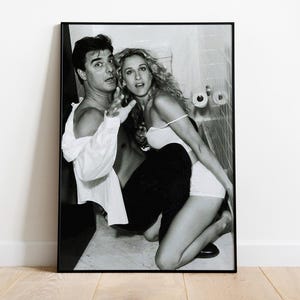 May include: A black and white framed photographic print featuring a man in an unbuttoned white shirt and a woman in a light-colored top and shorts, both positioned in a bathroom setting with tiled walls and a toilet paper roll visible in the background.
